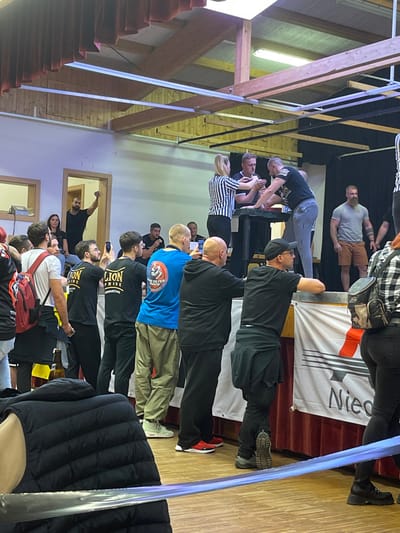 This image depicts an indoor arm wrestling competition taking place on a raised wooden stage within a hall, identified as being in Reisenberg, Austria.
On the stage, a female referee wearing a black and white striped shirt oversees an arm wrestling match between two male competitors. One male competitor, in a dark polo shirt, has his left arm on the arm wrestling table, opposing a second male competitor in a black short-sleeved t-shirt and light gray athletic pants, whose right arm is on the table. Both men are gripping each other's hands, positioned for the match. To the immediate right of the stage, a bearded man in a light gray t-shirt and brown shorts stands with his hands clasped in front of him. A second male figure is partially visible further to the right on the stage. Beneath the stage, a banner displays white text, partially visible as "Niedero," against a dark background with a red graphic.
A diverse audience, predominantly male, stands on the wooden floor observing the event. Many individuals in the midground are holding smartphones, seemingly recording or photographing the match. Notable individuals include: a man in a black "LION PRIDE ARM WRESTLING" t-shirt, another man in a blue t-shirt with a circular crest on the back, and a man in a black cap and t-shirt leaning on the stage banner. To the far left, in an open doorway, a man in a black t-shirt stands with his right fist raised in the air. In the immediate foreground, a person wearing a black puffer vest is partially visible from the back.
The venue features light-colored walls, exposed wooden beams across the ceiling, and fluorescent lighting fixtures. Red drapes are visible below the stage banner. The floor is made of wood.