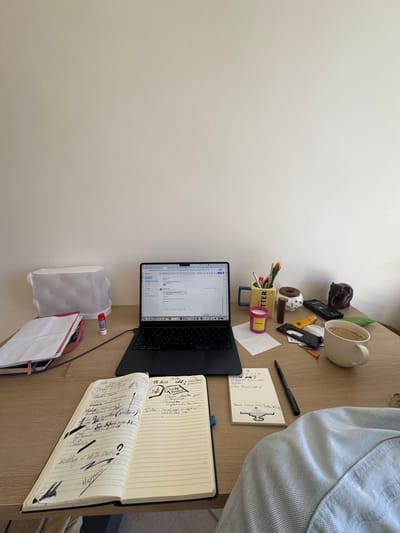 The scene depicts a light-colored wooden desk situated against a plain, off-white wall in a room within the Anavissos Municipal Unit, Greece. A dark-colored laptop computer is open and centrally positioned on the desk, its screen displaying an email client interface. A charging cable extends from the left side of the laptop.

To the left of the laptop, a stack of notebooks with a pink or red cover is visible, next to a white rectangular package of paper products wrapped in clear plastic and a white glue stick.

To the right of the laptop, a yellow mug holding various pens and pencils is present. A small pink cylindrical candle labeled "DAISY" rests on a crumpled white paper. Further right are a small brown donut-shaped decorative item, an irregularly shaped dark brown object, a black remote control, and an assortment of smaller items including orange plastic pieces and a yellow tube. An empty, light-colored ribbed ceramic mug, showing signs of residual liquid, lies on its side next to a green leaf-shaped object.

In the foreground, an open, dark-covered lined notebook is visible, filled with extensive handwritten notes, sketches, and crossed-out text on both pages. To its right, a smaller white lined notepad contains additional handwritten notes, including "PENA 2024" and a stylized drawing. A black pen rests between the notebook and the notepad. The lower right corner of the image shows a portion of a light blue denim-textured garment.