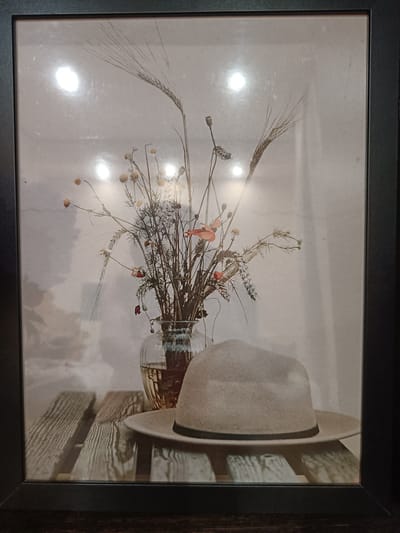 This is a framed photograph depicting a still life arrangement. The photograph is of a hat placed on a wooden surface, with a vase of dried flowers and wheat stalks behind it. The hat is light-colored with a dark band. The vase is glass and contains a bouquet of dried flowers, some of which appear to be poppies and small daisies, along with dried wheat stalks. The background of the photograph is a light, textured wall with circular reflections, possibly from studio lighting. The wooden surface resembles a rustic table or planks. The overall mood is subdued and artistic. The image does not provide any specific information about its location in Vichuga, Russia, nor does it contain any people or actions.