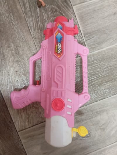 A pink toy water gun is lying on a wooden floor. The toy gun is made of plastic and has red accents. It has a handle, a trigger, and a barrel. There is a white reservoir at the bottom of the gun with a yellow cap. The gun is decorated with a shield-shaped emblem that reads "MX POWER" in red and blue letters. The wooden floor is made of planks of varying shades of brown and gray. The scene appears to be indoors.