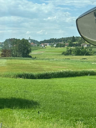 The image shows a rural landscape with rolling green fields under a cloudy sky. In the distance, a village with a church steeple is visible, nestled among trees on a gentle slope. The foreground is dominated by lush green grass, with a shadowed area suggesting the presence of an object or structure not fully within the frame. A car's side mirror is partially visible in the upper right corner. The scene depicts a tranquil, pastoral setting likely in the vicinity of Lindau (Bodensee), Germany.