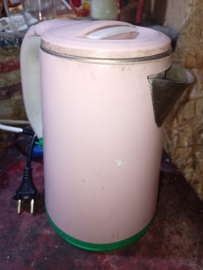 A pink electric kettle with a green base and a metal spout sits on a textured, dark red surface. A black electrical plug with two prongs is visible to the left of the kettle. The background appears to be a wooden wall. The scene is set in a cluttered environment, possibly a kitchen or workshop, in Zeravshan-2, Tajikistan.