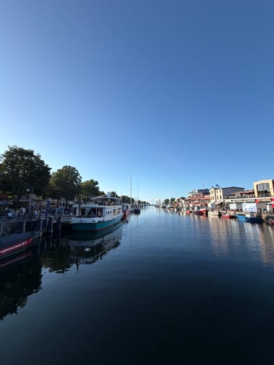The image depicts a canal in Rostock, Germany, likely in the city harbor area, lined with boats and buildings. A clear blue sky dominates the upper portion of the frame. On the left side, a docked boat with red lettering that reads "rophenschutz M-V" is visible. Next to it, a larger white and teal boat is moored, with people seated at tables on the adjacent pier, suggesting an outdoor seating area for a restaurant or cafe. Further along the pier, more boats are docked, and buildings with multiple stories, some featuring balconies and awnings, line the canal. The water reflects the sky and the surrounding structures, creating a peaceful scene. Several other boats, including sailboats, are visible further down the canal, extending into the distance. The overall impression is of a lively, scenic waterfront in a German coastal city.