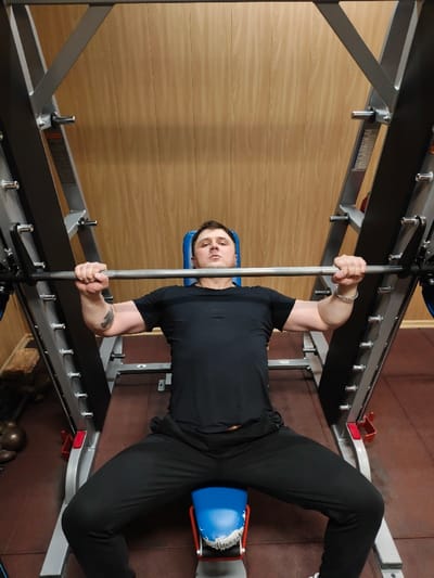 An adult male is performing a bench press exercise within a gym located in the city of Im. Gastello, Russia. The male, Caucasian with short dark hair, is positioned supine on an adjustable workout bench with a blue seat and backrest, which is integrated into a Smith machine. He wears a black short-sleeved athletic shirt with two horizontal stripes on the chest and black athletic pants. His hands grip a silver barbell, which is part of the Smith machine, in an overhand grip, with his arms slightly bent, indicating a paused or mid-lift position. A dark-colored circular tattoo is visible on his left bicep, and a silver-colored chain bracelet is on his right wrist. The Smith machine frame is metallic gray with numerous adjustable pins and weight plate holders; a dark blue weight plate is partially visible on the right side of the barbell. The floor is covered with dark reddish-brown rubber gym mats. The background wall features light brown wooden paneling. Additional gym equipment, including what appears to be dumbbells or kettlebells, is dimly visible in the far left background.