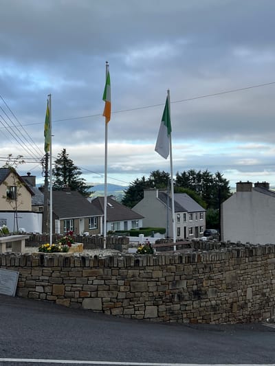 The image depicts a street scene in Mountcharles, Ireland. Three flags are prominently displayed on flagpoles. The top flag is a tricolor of green, white, and orange, representing the Irish flag. Below it, another flag also appears to be the Irish flag. The lowest flag is partially visible and seems to have green and yellow elements.
The flags are set against a backdrop of houses and a distinctive stone wall. The stone wall, constructed from irregularly shaped stones, forms a boundary along the right side of the road. Behind the wall and houses, trees and distant hills can be seen, suggesting a somewhat rural or semi-rural setting. The sky is overcast with clouds, indicating a possibly cloudy or moody day. There are no people visible in the image.
The scene appears to be a static street view, possibly a memorial area or a public space where the flags are flown. The presence of the Irish flags suggests a patriotic or nationalistic sentiment. The time of day is difficult to determine precisely due to the overcast conditions, but it is likely daytime. There is a small plaque or sign visible at the base of the stone wall, but the text on it is not legible. The overall impression is a quiet, somewhat somber, but visually rich depiction of a location in Ireland.