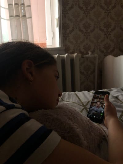 The media file shows a close-up of a young person lying down and holding a smartphone. The person's face is partially obscured, but they appear to be looking at the screen. The phone screen displays a video of a person with glasses and a surprised expression. The background suggests an indoor setting with patterned wallpaper and a window with blinds. The lighting is dim, indicating it might be evening or a dimly lit room. The person is wearing a striped shirt. The overall mood is relaxed and engaged with the content on the phone.

The scene takes place in Calfa, Moldova.
