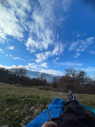 A serene landscape showcases a vibrant blue sky adorned with wispy white clouds, contrasted by a grassy terrain dotted with trees. A distant mountain range is partially obscured by mist. In the foreground, a person reclines on a blue cloth, their leg and sneaker visible. A slender stick is held casually, hinting at relaxation amidst nature's beauty.