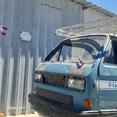 An aging, light blue Volkswagen Transporter van, with visible rust and damage, is parked in front of a corrugated metal wall. The van's front end is partially disassembled, with the grille and bumper removed. A ladder rack is mounted on the roof of the van. A small, white sign with Arabic text is attached to the metal wall to the left of the van. The ground is covered in sand and debris. The scene is set against a clear blue sky. The watermark "Galaxy A14" is present in the bottom left corner of the image.