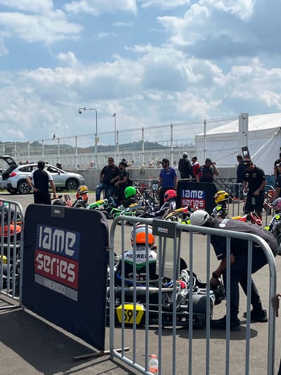 The image shows a kart racing event, specifically the IAME Series Mexico, taking place in Amozoc de Mota, Mexico. Multiple karts are lined up behind a metal barrier with the IAME Series Mexico banner attached. People are seen working on and around the karts, including mechanics and team members. The environment includes an outdoor racing venue with a partly cloudy sky overhead, as well as light poles and some surrounding buildings in the distance. The location is likely a karting track in the city of Amozoc de Mota.