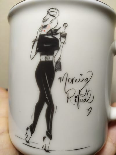 This is a close-up, rotated photograph of a white ceramic mug. The mug features a black graphic illustration of a woman in a fashionable outfit, holding a cup to her lips. The woman is depicted with red blush on her cheeks and a slight smile. She is wearing a black, sleeveless top with a defined waist cinched by a belt, and black pants with high-heeled shoes. A black bow is tied at her neck.

Above the illustration, the words "Morning Ritual" are written in elegant, cursive black script. A small, stylized heart is placed to the right of the text. The illustration and text are printed on the exterior surface of the mug. The background is a neutral, light-colored surface. The lighting appears to be soft and ambient. The image is taken from a slightly upward angle, showing the curve of the mug. The location context of Bilajari, Azerbaijan, does not provide any visual cues within the image itself.