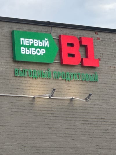 A sign for a B1 grocery store is mounted on a brick wall. The sign is green and displays the words "ПЕРВЫЙ ВЫБОР" in white. To the right of this sign are large red letters that spell out "B1". Below the main sign, smaller green letters on the brick wall spell out "ВЫГОДНЫЙ ПРОДУКТОВЫЙ". There is a security camera mounted on the wall below the signs. The sky is overcast. The store is located in Dedovsk, Russia.