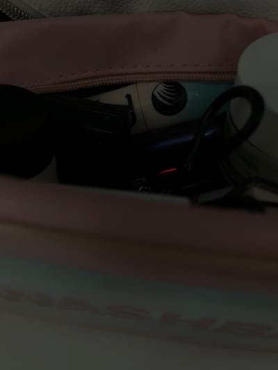 This is a close-up, low-angle, indoor, eye-level shot of the contents of a pink and white cosmetic bag. The bag is partially open, revealing various beauty products. On the left, a dark, cylindrical object, possibly a makeup brush or applicator, is visible. In the center, a tube with a white and teal design is partially obscured by a dark object. The tube has a circular logo with stylized eyelashes. To the right, a white jar with a green label is partially visible, with text that appears to include "MBER CREAM". The overall lighting is dim, with a red glow emanating from a small object in the lower center of the frame. The background is blurred, showing the textured interior of the cosmetic bag. There is no indication of the location being Pănășești, Moldova.