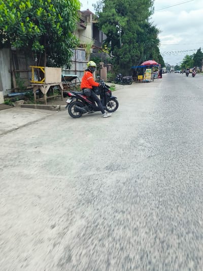 The image shows a man in a red jumpsuit and a yellow helmet riding a black motorcycle on a paved road in Kecamatan Dayeuhkolot, Indonesia. The road is empty except for two other motorbikes in the distance. The man is driving away from the camera towards a collection of vendors selling food on the right side of the road. The vendors have an awning above their stand and a sign that reads "Bakso." There are houses and trees along the sides of the road, and the sky is cloudy.