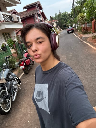 A person wearing headphones and a grey Nike t-shirt is taking a selfie on a street next to two parked motorcycles, near a red car on a road, with buildings and trees in the background. A dog is on the road in the distance. The location is Marra, India.