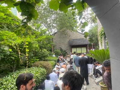 A gathering of men, many wearing traditional Muslim attire, are seated or standing outdoors in Qinhuai District, China. The group is situated on a stone-paved area, bordered by greenery, including trees, bushes, and potted plants. The architectural style of the surrounding buildings includes a grey brick building with a traditional tiled roof. A grey stone archway frames part of the scene.