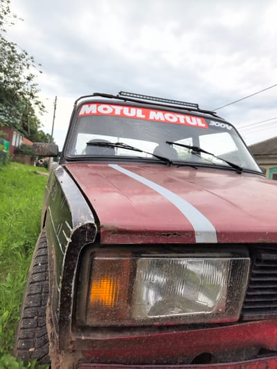 A close-up, low-angle exterior shot of the front of a red car shows a muddy, worn condition. The car is parked outdoors, next to a grassy area and a fence, suggesting a rural or suburban setting, possibly in the village of Koyanovo, Russia, given the context. The car's windshield has a red banner with white lettering that reads "MOTUL" and "300V." The windshield is cracked. A light bar is mounted on the roof of the car. The right front headlight is illuminated with an orange tint, and the left headlight is visible but appears dull. The front fender is dark colored and appears damaged. The car's tire is visible on the left side of the frame, covered in mud. In the background, there is a house with a gray roof and some trees. The sky is overcast.