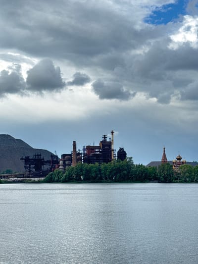 This is a vertical, eye-level, medium shot of an industrial landscape and water. In the foreground, a wide expanse of rippling water occupies the lower half of the frame, reflecting the overcast sky. Behind the water, a line of dense green trees forms a natural barrier. Beyond the trees, on the left, is a large, dark industrial complex with tall smokestacks and intricate metal structures, possibly a blast furnace. To the right of the industrial complex, nestled amongst the trees, are two buildings with red and gold domes, characteristic of Russian Orthodox churches. The sky above is filled with dramatic, heavy gray clouds, with a small patch of blue sky visible in the upper right quadrant. The overall impression is one of industrial might juxtaposed with natural elements and religious architecture, under a moody, overcast sky. This scene is located in the city of Satka, Russia.