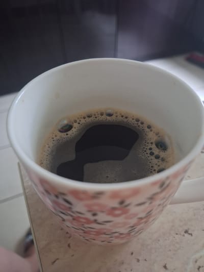 A close-up, eye-level shot captures a white ceramic mug filled with dark coffee. The mug has a pink floral pattern on the exterior. Bubbles on the surface of the coffee create iridescent effects. The mug rests on a light-colored surface, possibly a countertop with a tiled pattern. In the background, a dark, out-of-focus surface, likely a cabinet or wall, is visible. The image does not contain any discernible information to place it in the city of Mińsk Mazowiecki, Poland.