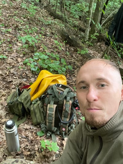 A man with short hair, a light complexion, and a goatee is taking a selfie in a forest. He is wearing a green hooded jacket. To his left, there are several pieces of camping gear, including a large green backpack, a yellow tarp, and a metal thermos. The ground is covered in fallen leaves and vegetation. The background shows trees and more foliage. The image does not contain any elements that would indicate it is located in the city of Nikolayevka, Russia.