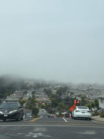The image shows a residential street in San Bruno, California, United States of America, on a foggy day. A black Toyota Sienna minivan is parked on the left side of the street, facing uphill. A white Tesla Model 3 is parked on the right side of the street, also facing uphill. There are construction signs and orange traffic cones on the side of the road, indicating road work. The street is lined with houses and trees, and the city extends into the distance, shrouded in fog. The sky is overcast and light gray.