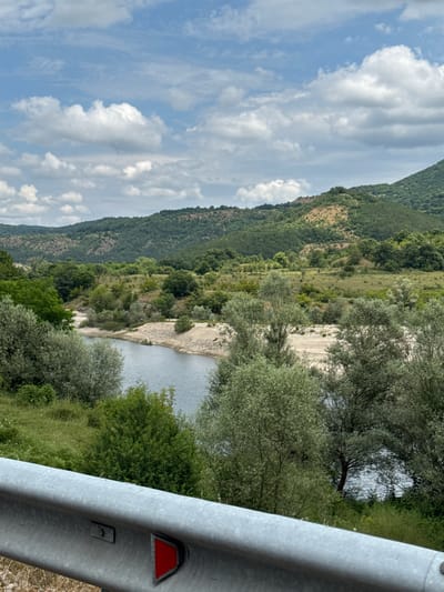 This is a landscape photograph taken outdoors, featuring a natural scene with a river winding through a valley. The image is oriented vertically. In the foreground, a metal guardrail runs along the right side of the frame. The guardrail is silver and has a red triangular reflector attached to it.
Beyond the guardrail, lush green trees and vegetation dominate the midground. A body of water, likely a river, flows through the scene, with its surface reflecting the sky. The riverbanks are a mix of sand and pebbles. In the background, rolling hills covered in dense green trees rise up, suggesting a mountainous or hilly terrain. The sky is partly cloudy, with scattered white clouds against a blue background.
The time of day appears to be daytime, given the brightness and cloud formations. The weather seems pleasant, possibly warm and sunny, judging by the vibrant green of the foliage and the clear blue sky. There are no people visible in the image. The overall impression is one of natural beauty and tranquility, a scenic view of a rural or wilderness area. The presence of the guardrail suggests the photo may have been taken from a road overlooking the landscape. While the exact location is not explicitly indicated by text in the image, the context provided places it in Bregovo, Bulgaria.