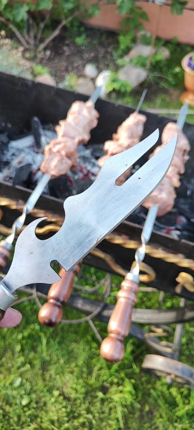 In an outdoor setting, possibly a backyard, a person is holding a metal skewer with raw meat on it, with a barbecue grill in the background. The skewer has a unique, curved metal handle with a bottle opener-like feature. The barbecue grill contains hot coals and several more skewers of meat. The ground is covered in green grass. The image appears to be taken during daylight. There are no visible landmarks or indicators to suggest the location is specifically in Лёзненскі сельскі Савет, Belarus.