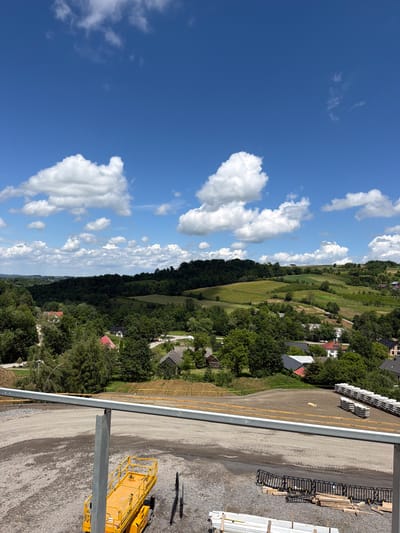 In Jodłownik, Poland, an elevated view captures a construction site in the foreground with a yellow scissor lift and stacked building materials. A gravel ground surface is visible, bordered by a metal railing. Beyond the construction area, a verdant landscape unfolds with rolling hills covered in dense green trees. Scattered amongst the trees are residential buildings with red and grey roofs, indicating a small village. The sky above is a clear, bright blue with scattered white cumulus clouds. The overall scene depicts a sunny day with ongoing development within a rural setting.