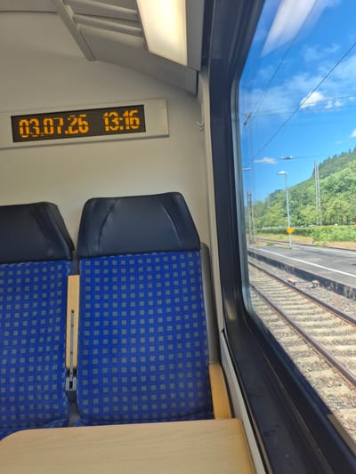 The image shows the interior of a train carriage with two blue and grey patterned seats visible. To the left of the seats, an orange digital display shows the date "03.07.26" and the time "13:16". Through the window on the right, railway tracks are visible, alongside a platform and a hillside covered in trees. The sky is blue with some white clouds. The train appears to be stopped at a station in Kobern-Gondorf, Germany, as suggested by the presence of the station and the surrounding landscape.