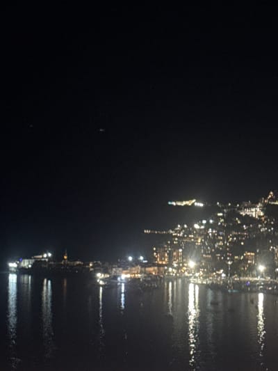 The image is a nighttime, slightly blurry photograph of a coastal town. The foreground is dominated by dark water, reflecting the lights of the town in streaks of white and yellow. The town itself is built on a hillside and is illuminated by numerous lights, giving it a warm glow. Buildings, possibly houses and hotels, are visible with many illuminated windows. There are also streetlights or lamp posts. In the distance, across the water, is a densely lit area that appears to be a more significant part of the town. The sky above is dark and clear, with a few faint specks of light, possibly stars or distant aircraft. There are no visible people or specific identifiable structures that would allow for a definitive location identification. However, the style of the architecture and the coastal setting could be consistent with towns found in the Mediterranean region.