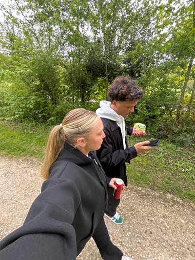 The image shows two individuals walking on a path in an outdoor environment, presumably in Noisiel, France, based on provided contextual data. The person in the foreground, with blond hair in a ponytail and wearing a black jacket, is partially visible. The second person, positioned slightly behind, has curly brown hair, is wearing a black jacket over a white hooded sweatshirt, and is holding a smartphone in one hand and a package of sweets in the other. This individual also has a can of what appears to be soda in their other hand. They are walking on a gravel path, surrounded by green vegetation including trees and bushes, indicating a park or wooded area. The scene appears to be captured at eye level.