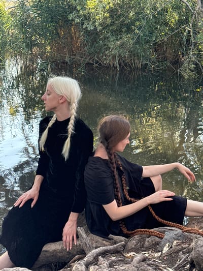The image shows two individuals from the back, sitting on rocks by a body of water. The person on the left has blonde hair in braids, and the person on the right has brown hair in a braid. Both are wearing dark clothing. The environment appears to be natural, with lush green foliage and reeds along the edge of the water, and the reflection of trees is visible on the water's surface. The scene suggests a peaceful and contemplative setting, possibly near the shores of the Sea of Galilee, which is a significant geographical feature in the city of Katzrin, Israel.
