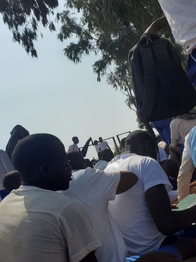 The image depicts an outdoor gathering of people, likely students, in Barakin Rafin Gora, Nigeria. In the foreground, several individuals are seen from behind, wearing white t-shirts and blue shorts. One person in the center is wearing a white t-shirt and appears to be looking to their right. To the left, a person's head and shoulder are visible, wearing a light-colored t-shirt.

In the middle ground, a person in a white t-shirt is visible from the side, and behind them, another person in a white t-shirt and blue shorts is seated. Further back, a group of individuals in white t-shirts are standing, some holding dark flags. To the left, a person wearing a dark head covering is visible. A black backpack is being held by a person in the upper right quadrant of the image, who is wearing a white t-shirt and blue shorts.

The background consists of a bright, overcast sky and the branches of eucalyptus trees, characteristic of the Nigerian landscape. A metal structure, possibly part of a bleacher or seating area, is visible in the middle ground. The ground appears to be reddish-brown earth. The overall impression is of a community event or gathering in an open-air setting.
