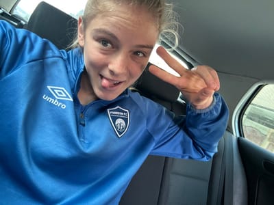 A young individual, identified as female, is depicted seated in the rear passenger compartment of a vehicle, in the city of Borgo San Dalmazzo, Italy. She is looking directly at the camera with a playful expression, her tongue slightly extended. Her light-colored hair is pulled back. She wears a long-sleeved, bright blue athletic quarter-zip top. A white Umbro logo is visible on the left chest. On the right chest, a shield-shaped patch features "FREEDOM FC" text above a mountain graphic, with "Libertà fare andaré oltre" below. Her left hand is raised, forming a "peace" sign (V-sign) with her index and middle fingers extended. The vehicle's interior shows dark grey fabric seats and a light grey headliner. A blurred exterior view is visible through the passenger side window.