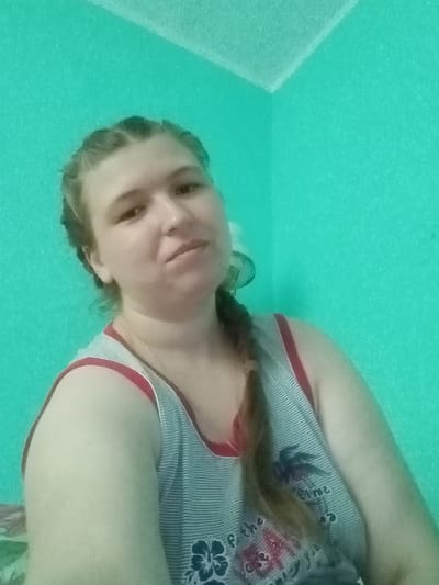 A young woman is pictured from the chest up, facing the camera at a slight angle. She has light skin and light brown hair braided down her right side. She is wearing a gray and white striped tank top with a red trim. The background is a bright teal wall, with a hint of another wall at a corner to the left. The lighting suggests an indoor setting, likely a room. The image quality is somewhat grainy. The provided location, Kitskany, Moldova, cannot be visually confirmed or denied from the image alone.