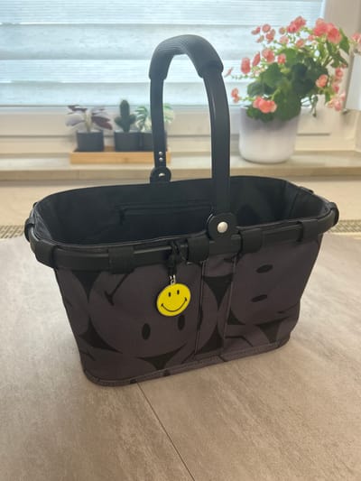 A gray tote bag with a smiley face pattern sits on a neutral-toned tiled surface. The bag has black handles and a black interior with a zipper pocket. A yellow smiley face charm hangs from one of the handles. In the blurred background, a window with horizontal blinds is visible, with several potted plants on the windowsill, including a cactus and a plant with purple leaves. To the right, a pot with pink flowers is also visible. The scene is indoors, likely in a domestic setting in Velbert, Germany. There are no people present in the image.