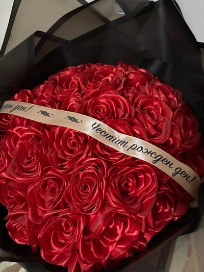 A bouquet of red fabric roses is presented in a black wrapping. A light-colored ribbon is draped across the roses with text in a cursive font. The text reads "Честит рожден ден!" which translates to "Happy Birthday!" The background is dark and out of focus, suggesting an indoor setting. There are no people or animals present in the image. The image is a close-up shot of the bouquet.