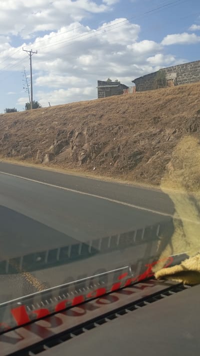 A roadside view with a two-lane asphalt road in the foreground. Above the road is a grassy embankment, leading to a flat area with two small buildings, one appearing brick and the other made of corrugated metal. A utility pole and wires are visible against a sky with scattered clouds. The perspective is from inside a vehicle, with parts of the dashboard visible.