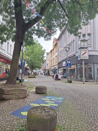 The image shows a cobblestone street in Bochum, Germany, lined with shops and trees. Colorful flags are strung across the street, and a large tree with a rough trunk stands prominently in the foreground. There are several concrete planters and large, rounded stone bollards on the sidewalk. A clock is visible on a pole, along with signs indicating "Zu vermieten" (For rent) and "KIND" (Child). In the distance, people are walking and sitting, suggesting a public thoroughfare. The buildings on either side of the street have a mix of architectural styles. The overall scene depicts a typical European street with commercial activity and decorative elements.