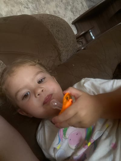 In this vertically oriented, close-up image, a young child is the central subject. The child, with fair hair and large brown eyes, is lying on their side and appears to be holding an orange pacifier in their mouth. The child's face is turned towards the camera, and their expression is calm. The child is wearing a white t-shirt adorned with a pastel-colored unicorn design, featuring pink, purple, green, and yellow accents.
The child is positioned on a textured, dark brown surface that resembles a couch or upholstered furniture. To the left of the child, a darker, wooden piece of furniture is visible, possibly a table or cabinet, with a metallic object protruding from it. The background is mostly dark and out of focus, suggesting an indoor setting. The lighting appears to be dim, possibly indicating an evening or indoor time of day.
The image was captured in Pesnevo, Russia. There is no visible text in the image. The overall mood is intimate and captures a private moment with the child.