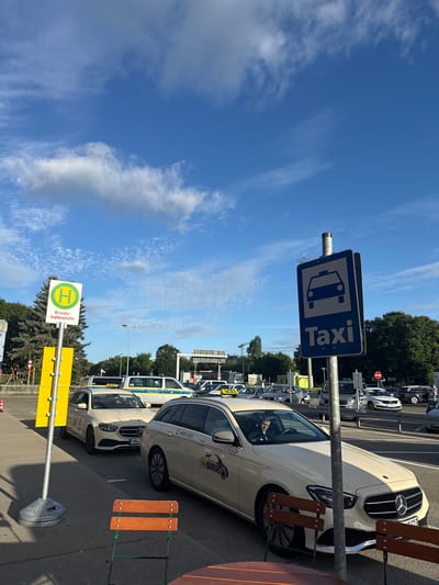 The image captures a bright, sunny day with a clear blue sky dotted with fluffy white clouds, at the Künersberg, Germany, taxi stand. The scene is dominated by several light-colored taxis, likely Mercedes-Benz sedans, lined up in a queue. One taxi in the foreground displays "TAXI FEKLER" on its side and has the phone number "0331-2323" visible. Another taxi further back has a driver behind the wheel. A police van, marked with "POLIZEI," is also present in the lineup.

To the left, a green and white bus stop sign with a yellow "H" symbol and the German text "Ersatz-haltestelle" (temporary stop) stands prominently. Behind the bus stop sign, a dense line of evergreen trees forms a backdrop. In the middle ground, a traffic signal and other vehicles are visible, suggesting a road intersection or a busy transportation hub. In the foreground, two wooden chairs with metal frames are set up, hinting at a waiting area or an outdoor seating space. The overall impression is one of an organized taxi stand in an urban or semi-urban setting on a pleasant day.