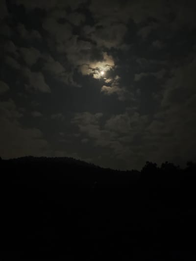 The scene depicts a nighttime landscape in La Marquesa, Mexico. The sky occupies the upper two-thirds of the image, presenting numerous scattered cumulus clouds illuminated by a bright, full moon located slightly right of the upper center. The moonlight creates varying shades of gray within the clouds, contrasting with the dark portions of the sky. The lower third of the image consists of a completely dark, undulating silhouette of a forested hillside or mountain range, devoid of specific detail due to the lack of light. The immediate foreground is entirely obscured by darkness. No people, man-made objects, actions, or interactions are discernible.