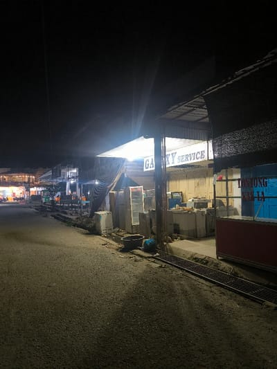 This image depicts a nighttime street scene in Manetunong, Indonesia. An asphalt road occupies the foreground and extends into the distance on the left. Alongside the road, a sidewalk features a metal-grated drainage ditch. The primary subject is an open-front commercial establishment located in the right-midground, prominently illuminated by an overhead light and a sign that reads "GALAXY SERVICE." This shop contains and is fronted by multiple large appliances, including refrigerators and washing machines. To the immediate right, a partially visible stall features a red counter and a blue section with text that appears to include "LONTONG KAK LA." Further down the street on the left, a row of other commercial buildings, dimly lit with various internal and external light sources, is discernible. No individuals are present in the visible scene.