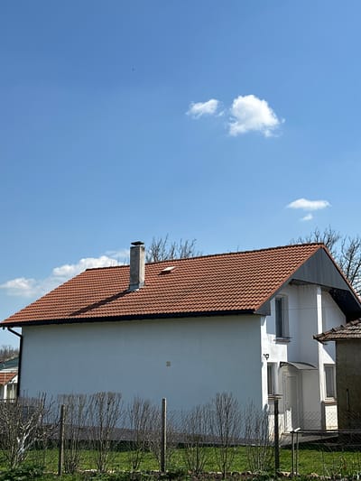 The image shows a two-story house in Balkantsi, Bulgaria. The house is white with a red-tiled roof and a chimney. A narrow fence runs along the side of the house, separating it from a grassy area with small bushes. The sky is blue with a few scattered clouds. There are no people visible in the image.