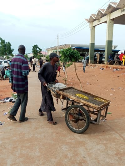 The image shows a street scene in Gombe, Nigeria, under an overcast sky. In the foreground, a man in a dark robe is pushing a wooden cart with a single wheel. The cart contains some white bags and what appears to be yellow food items. To the left of the cart pusher, a man in a purple and grey striped shirt and dark pants walks away from the camera. Behind them, other individuals are visible, some standing and some sitting. Several vehicles, including a white van and other cars, are parked in the background. A large, architectural structure with zig-zagging decorative elements and supporting pillars, possibly an entrance or a covered market area, is visible on the right side of the image, with text that appears to say "KANO" and "AZARE". The ground is predominantly reddish-brown dirt, with scattered litter. Trees are visible on the left side of the image.