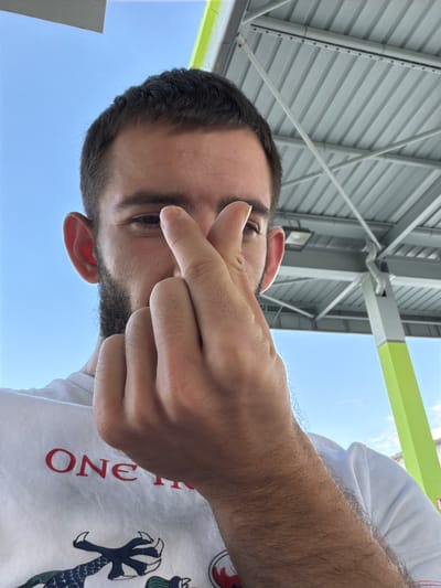 The image is taken from a low-angle perspective, looking up at a person standing outdoors. The person is light-skinned with dark hair and a beard. They are wearing a white t-shirt with a design that includes the word "ONE" in red letters and a graphic that appears to be a dragon claw. The person's hand is partially obscuring their face.
Above the person is a structural element composed of gray metal panels and beams, suggesting an awning or roof. There is a green stripe visible. Beyond the structure, the sky is visible and appears to be clear.
The location mentioned, Yoakim Gruevo, Bulgaria, cannot be confirmed or denied based on the image content.