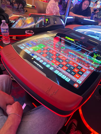 The image shows a person playing an automated roulette machine. The machine is branded "CRYSTAL". The screen displays the roulette layout with betting options and recent spin results. Casino lights are visible in the background along with other patrons. The scene appears to be inside a casino, given the roulette machine and gambling setting. Based on the information on the machine, the location is Hollywood Seminole Reservation, United States of America.