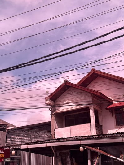 The image shows a street scene in Cotraya, Indonesia, dominated by a large building with a red-tiled roof. Overhead, a dense network of electrical wires crisscrosses the sky, which is filled with pinkish clouds, suggesting a sunset or sunrise. The building appears to be multi-story, with architectural details like columns and balconies visible. Below the main building, there are awnings and what seem to be storefronts or market stalls, featuring corrugated metal siding and visible signage, one of which reads "EL" and another "16.000". The overall impression is an urban environment with a mix of residential and commercial structures, characterized by the pervasive presence of overhead utility lines.