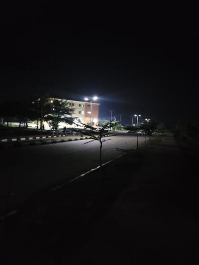 This nighttime image captures a street scene in Gaba, Nigeria. A multi-story building, featuring light-colored and reddish-brown facade sections, is visible in the background, illuminated by interior window lights and external fixtures. In the mid-ground, a paved road with a distinct curb and an unpaved dark shoulder extends from the foreground. Several small, sparsely foliated trees are planted at intervals along the road's edge. Multiple streetlights emit bright white light, illuminating sections of the road and the immediate vicinity. No individuals, animals, or vehicles are present within the frame.