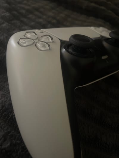 A close-up, angled shot focuses on the white and black buttons and joysticks of a video game controller, resting on a dark, textured surface. The controller, likely a PlayStation 5 DualSense, is oriented diagonally, with its rounded base filling the lower portion of the frame.
Four translucent, petal-shaped buttons are clearly visible in the lower left quadrant of the image. Above these, a black joystick with a textured grip is prominent. Another joystick is partially visible in the upper left. The controller's body transitions from white on the bottom to black on the top, with a thin white line separating the two sections. The background is a dark, fuzzy material, possibly a blanket or carpet, with subtle patterns.
The lighting appears to be dim, casting soft shadows and highlighting the reflective surfaces of the buttons and joysticks. No people are visible in the frame. The overall impression is one of a gaming accessory in a dimly lit, indoor setting. There is no visible text. The scene is purely an object-focused still life.