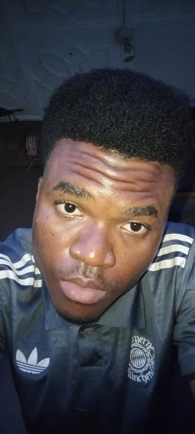 A close-up selfie shows a young Black man looking directly at the camera. He has short, dark, curly hair and light stubble on his upper lip and chin. His skin appears somewhat shiny, and his forehead has several horizontal creases.
He is wearing a dark blue polo shirt. On his left chest, there is a white Adidas Trefoil logo. On his right chest, there is a circular white emblem that reads "Bayern München" around a patterned center. White stripes are visible on the right shoulder area of his shirt.
The background is dark and appears to be indoors. A light-colored ceiling with a patterned texture is visible at the top, along with what looks like a ceiling light or fan fixture. The overall lighting suggests an indoor setting, possibly in the evening. The man's expression is neutral, looking intently towards the camera. No other specific activity is depicted besides posing for the photo.